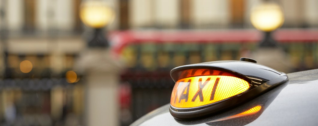 Picture shows a yellow light up sign with word 'TAXI' glowing in black on a black taxi - red bus in the background